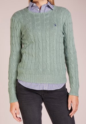 Femme portant un pull en laine torsadée vert clair sur une chemise à rayures bleues et blanches, avec un pantalon foncé, debout les bras détendus.