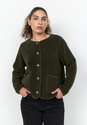 Femme aux cheveux bouclés portant une veste polaire vert foncé boutonnée avec poches avant et un pantalon noir, mains dans les poches, expression neutre.