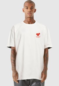 Weißes oversized T-Shirt aus Baumwolle, mit einem roten Herz-Graphic und Text im Bereich der linken Brust. Kurze Ärmel, lässige Passform.