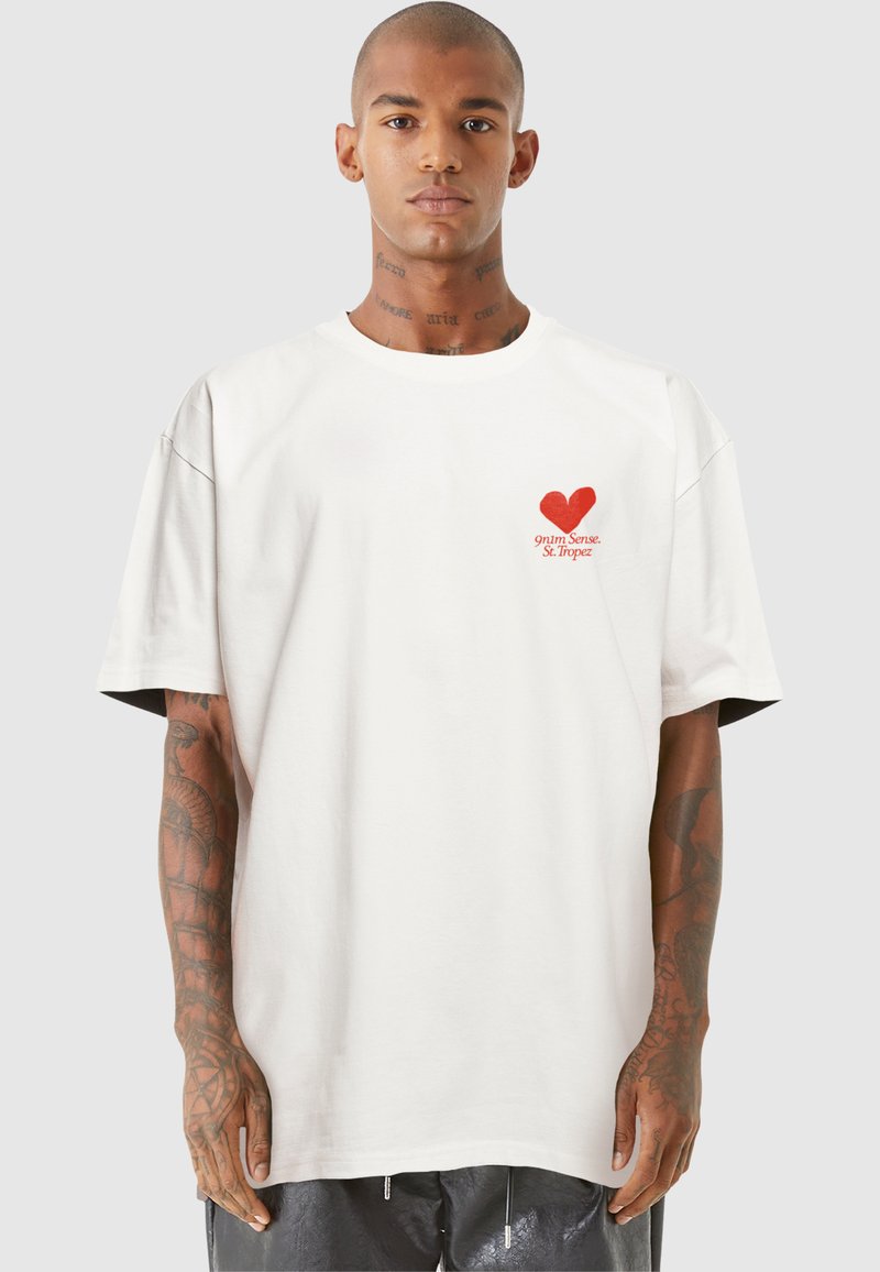 Weißes oversized T-Shirt aus Baumwolle, mit einem roten Herz-Graphic und Text im Bereich der linken Brust. Kurze Ärmel, lässige Passform.