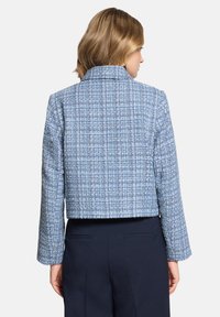 Femme portant une veste courte à carreaux bleus et blancs à manches longues et un pantalon bleu marine, vue de dos sur un fond uni.