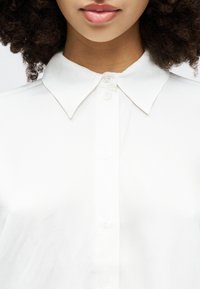 White button-up shirt with a classic collar, featuring a smooth texture and subtle sheen, shown at the collar and upper buttons.