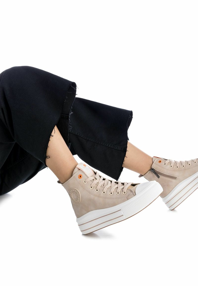 Zapatillas altas beige de ante texturizado, con suela de goma blanca, acentos naranjas y diseño de cordones. Combinadas con pantalones negros cortos.