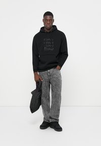 Hanorac negru cu text embosat, asortat cu blugi gri marmorați. Modelul ține o jacheta puffer neagră și poartă încălțăminte neagră. Fond simplu.