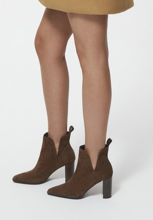 Brązowe zamszowe botki za kostkę z ostrym noskiem, wycięciami po bokach i słupkiem. Z tylu znajduje się pętla ułatwiająca zakładanie.