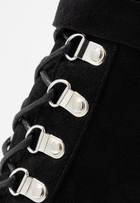 Botte en suede noire avec œillets en métal argenté et lacets noirs. Le design présente une texture lisse avec des détails de fixation bien marqués.