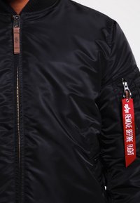Schwarze Bomberjacke aus Nylon mit Reißverschluss, geripptem Kragen und einem roten dekorativen Anhänger mit der Aufschrift "VOR DEM FLUG ENTFERNEN."