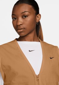 Smeđi zip-up prsluk preko bijele košulje dugih rukava, s crnim Nike logotipima na oba komada odjeće. Glatka tkanina s laganim dizajnom.