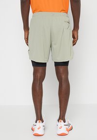 Pantalones cortos deportivos verde claro con cintura, bolsillo lateral y textura de malla, superpuestos sobre pantalones cortos de compresión negros; usados con zapatillas blancas y naranjas.