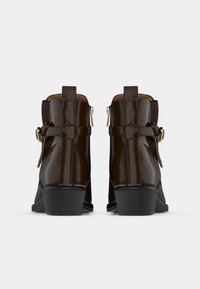 Par de botas de tobillo marrón oscuro con tacones bajos tipo bloque, cremalleras laterales y correas con hebilla ajustable, mostradas desde la parte trasera sobre un fondo blanco.