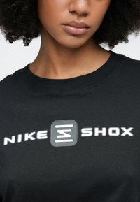 Μαύρο μπλουζάκι με το λογότυπο της Nike και τη λέξη "SHOX" σε λευκή και γκρι εκτύπωση. Το ύφασμα φαίνεται απαλό με κλασικό στρογγυλό λαιμόκοψη.
