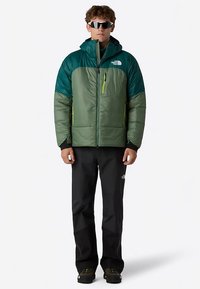 Groene en blauwgroene geïsoleerde jas met ritssluitingzakken en capuchon, gecombineerd met een zwarte broek. Voorzien van een opvallend logo en ritssluitaccenten in contrastkleur.