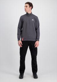 Šedá mikina na zips s vysokým golierom v kombinácii s čiernymi cargo nohavicami s viacerými vreckami. Červený štítok ako akcent na mikine.