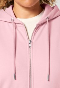 Felpa con zip rosa caratterizzata da una zip argento, cordini regolabili e una texture morbida. Presenta un cappuccio e un design semplice.