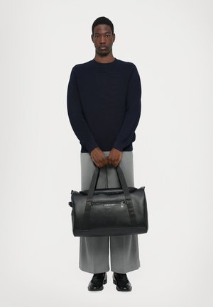 Black leather duffle bag with fabric handles, a zippered front pocket, and a textured finish, held by a person wearing gray pants and a navy sweater.
