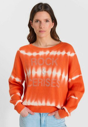 Vrouw met bruin haar, gekleed in een oranje-witte tie-dye trui met de tekst "ROCK & CERISES", handen in de zakken van blauwe spijkerbroek, neutrale achtergrond.