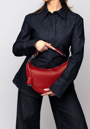 Red leather handbag with a curved shape, featuring a braided handle and a tag. The model wears a dark denim jacket and pants.