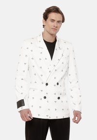 Mężczyzna w białym dwurzędowym blezerze z czarnym abstrakcyjnym wzorem, czarnej koszulce i czarnych spodniach, stojący na jednolitym tle.