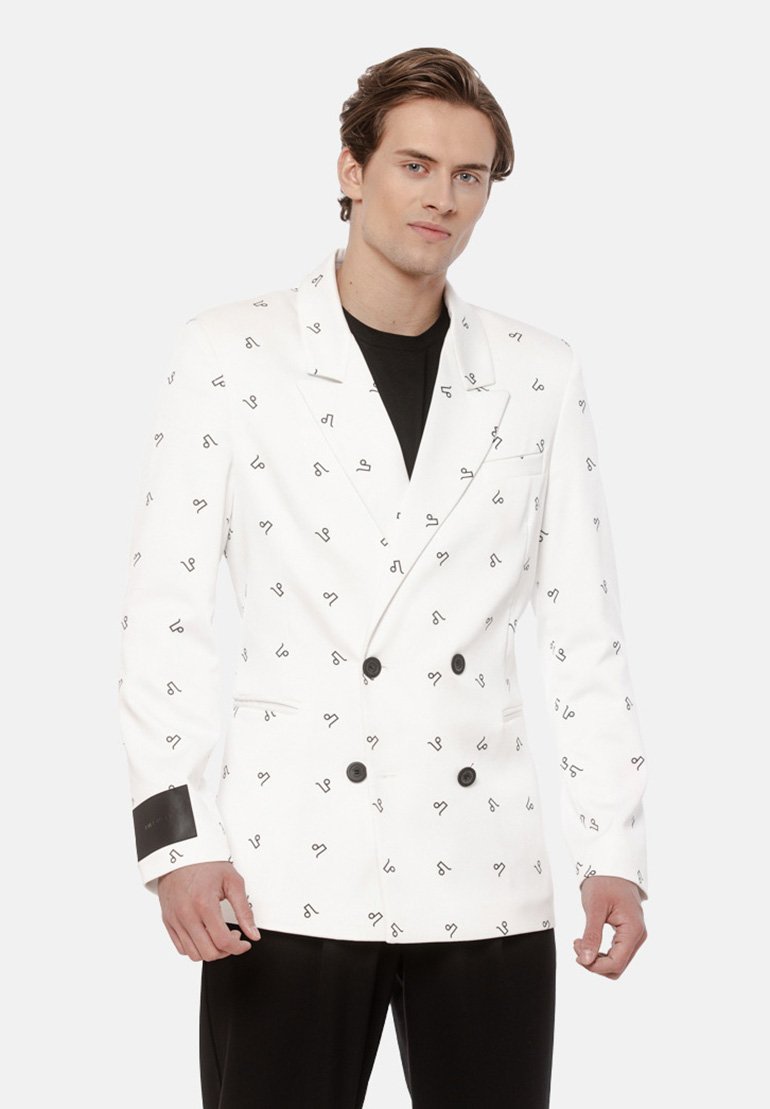 Mężczyzna w białym dwurzędowym blezerze z czarnym abstrakcyjnym wzorem, czarnej koszulce i czarnych spodniach, stojący na jednolitym tle.