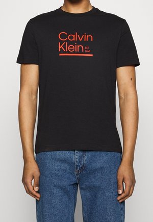 Mężczyzna ubrany w czarny T-shirt Calvin Klein z czerwonym napisem "Calvin Klein EST. 1968" w połączeniu z niebieskimi dżinsami, stojący na jednolitym tle.