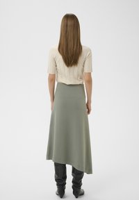 Beige geripptes Kurzarmoberteil kombiniert mit einem langen, olivgrünen asymmetrischen Rock und schwarzen kniehohen Stiefeln mit spitzer Form.