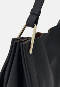 Gros plan d’un sac à main en cuir noir texturé avec des côtés plissés, une bandoulière réglable et des ferrures métalliques rectangulaires dorées sur fond blanc.