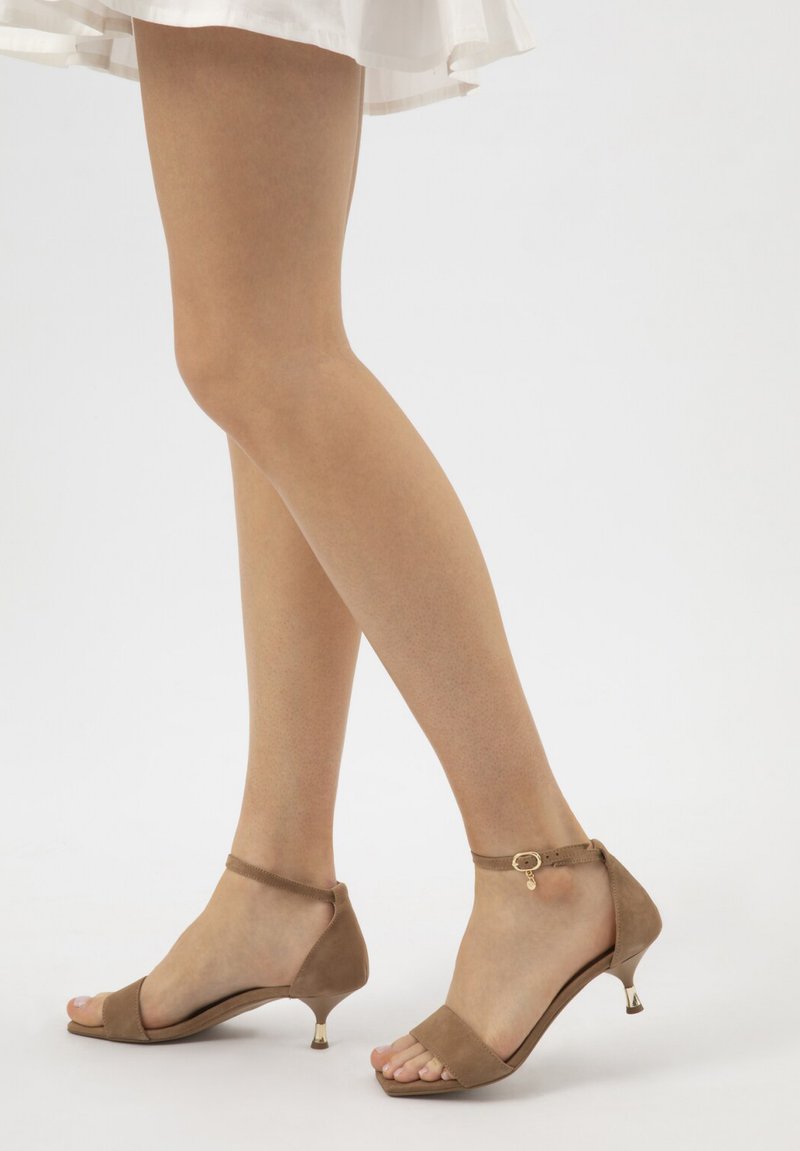 Jambes portant des sandales à talons plats avec bride à la cheville de couleur beige, assorties à une jupe blanche, debout devant un fond blanc uni.