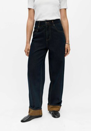 Personne portant un jean large foncé avec des revers retroussés, des chaussures noires et une chemise blanche à manches courtes, debout devant un fond blanc.