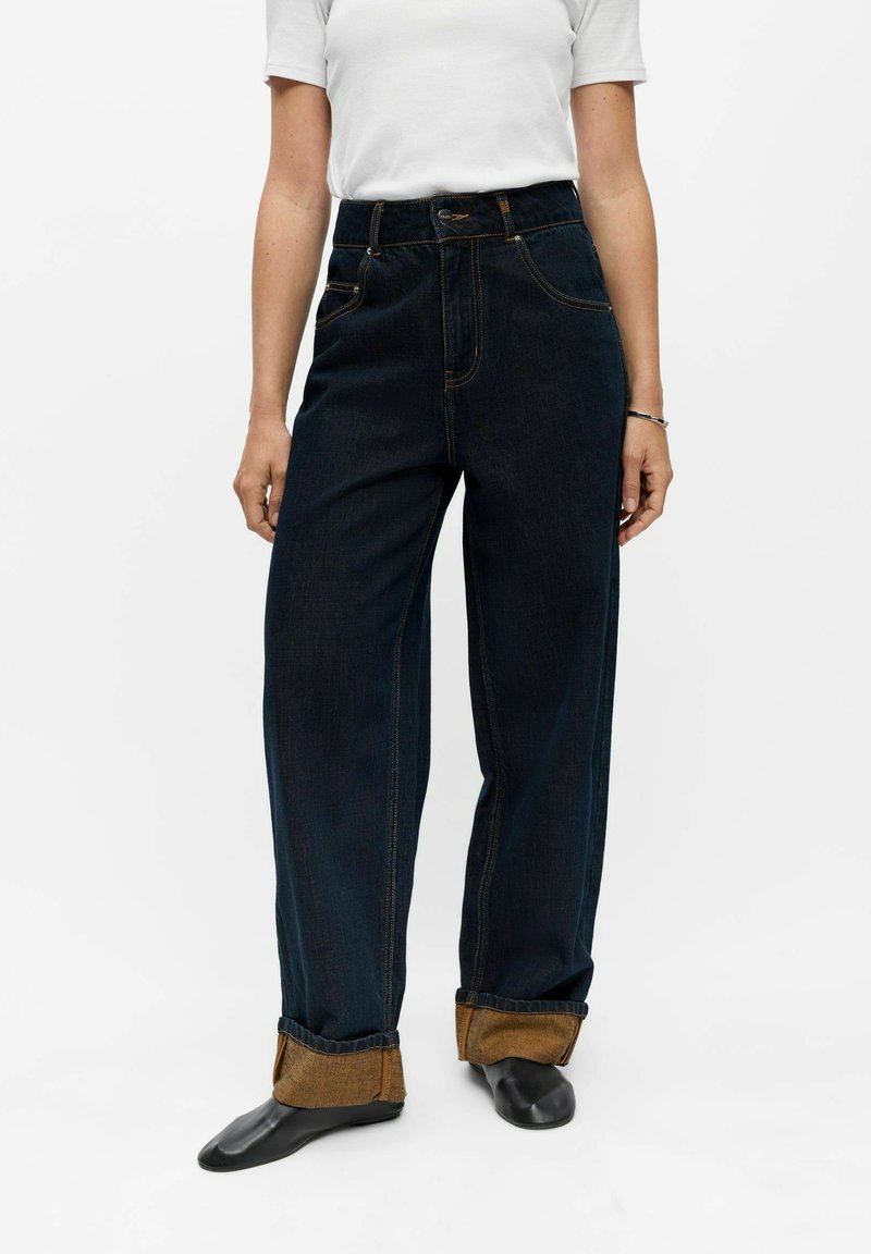 Personne portant un jean large foncé avec des revers retroussés, des chaussures noires et une chemise blanche à manches courtes, debout devant un fond blanc.