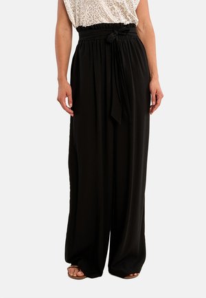 Femme portant un pantalon noir large à taille haute avec une ceinture nouée et un chemisier léger à imprimé léopard rentré, debout les mains détendues.