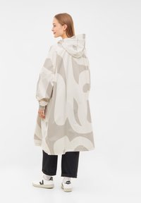 Lätt, huva regnjacka i krämfärgad med abstrakta beige mönster. Har elastiska manschetter och en avslappnad passform, kombinerad med svarta byxor och sneakers.