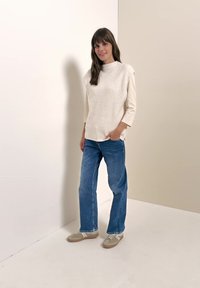 Femme debout contre un mur uni, portant un pull sans manches en tricot crème, une chemise à manches longues, un jean bleu et des baskets beiges, souriante avec une main dans la poche.
