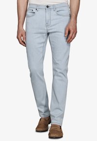 Hombre con vaqueros ajustados azul claro y mocasines de ante marrón, de pie con las manos relajadas a los lados.