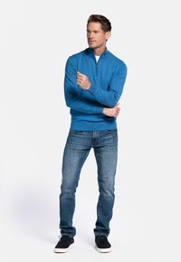Blauwe zip-up trui met lange mouwen, gestructureerde stof, gecombineerd met donkere spijkerbroeken en donkere schoenen, staand tegen een effen achtergrond.