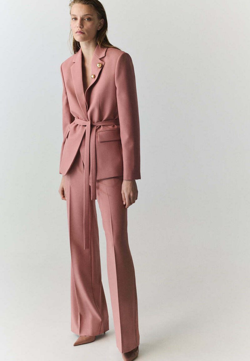 Femme debout portant un blazer ceinturé rose poudré avec des boutons dorés et un pantalon large assorti, sur un fond clair uni.
