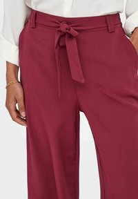 Pantalon large couleur bordeaux avec un tissu texturé, doté d'une ceinture nouée à la taille et de poches latérales. Associé à un chemisier blanc.