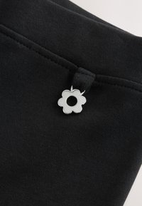 Nahaufnahme eines schwarzen Stoffs mit einem kleinen silberfarbenen, blumenförmigen Metallanhänger nahe einer Naht.