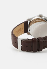 Bracelet de montre en cuir marron avec des coutures contrastées, fermoir en acier inoxydable couleur argent et logo de la marque gravé sur le côté intérieur.