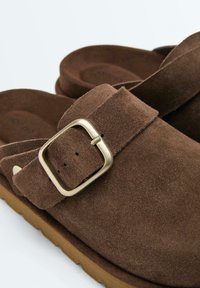 Bruine suède instapsandalen met verstelbare gouden gespbandjes en getextureerde lichtbruine rubberen zolen.
