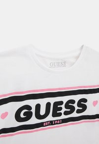 White organic cotton t-shirt with a crew neck, featuring black and pink horizontal stripes, large "GUESS" logo, and "EST. 1981" text below.