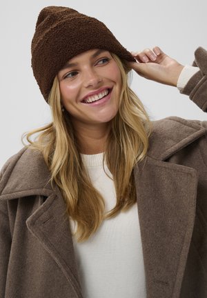 Bruine, textuur gebreide muts met een iets puntige top, gecombineerd met een beige trui en een bruine wollen jas, die warme, neutrale tinten laat zien.