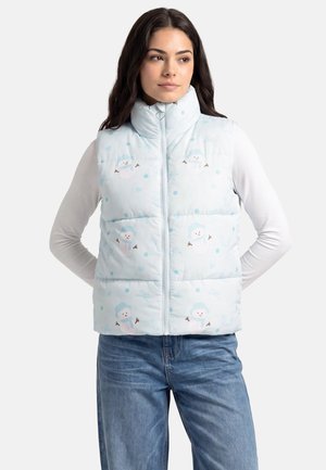 Frau trägt eine hellblaue Steppweste mit Schneemannmotiven, weißes Langarmshirt und blaue Jeans, steht mit den Händen hinter dem Rücken.