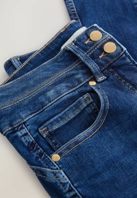 Mėlyni džinsai su aukštu liemeniu, turintys du auksinius mygtukus, kontrastingą siuvinėjimą ir priekinį kišenių detalę.