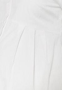 Vit blus med knappstängning, en plisserad design vid midjan och en slät, mjuk textur. Inga synliga mönster eller detaljer.