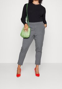 Pantalon gris ajusté avec une coupe slim, associé à un top noir à manches longues côtelé, des chaussures à talons hauts rouges et un sac à main tissé vert.