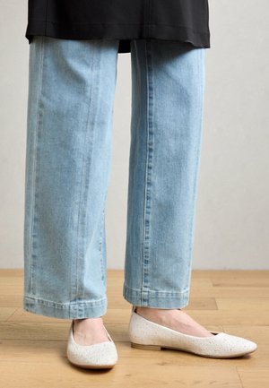 Untere Beine, die hellblaue Jeans mit weitem Bein und weiße strukturierte flache Schuhe tragen, stehen auf einem Holzboden.