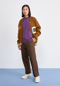 Bruine varsityjack met witte strepen, een logo en tekst, gedragen over een paarse t-shirt. Gecombineerd met bruine broek en crèmekleurige sneakers.