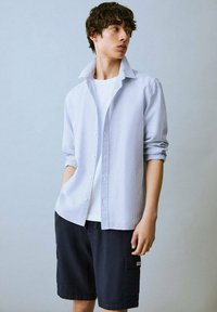 Camicia button-up a righe blu chiaro con colletto aperto, indossata sopra una maglietta bianca, abbinata a pantaloni corti blu navy scuri. Tessuto morbido, vestibilità comoda.