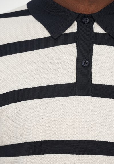 Kötött pólókrém színben fekete vízszintes csíkokkal, fekete gallérral és kétgombos gombolással. Texturált anyag légáteresztő szövettel.