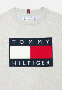 Hanorac gri cu un logo dreptunghiular bleumarin și roșu ce conține textul „TOMMY HILFIGER”. Guler striat cu detaliu interior cu dungi.